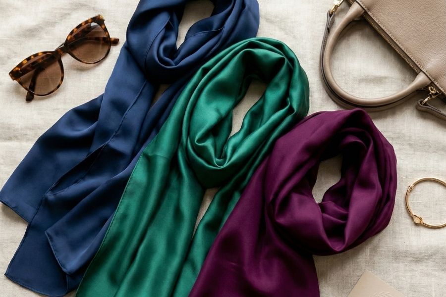 Three scarves in blue, green, and purple with a handbag and sunglasses on a light surface.